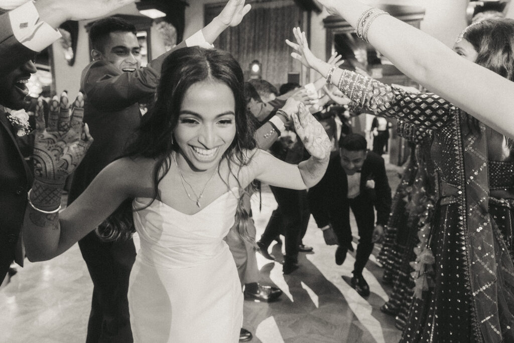 The bride, beaming with happiness, runs through a tunnel of arms created by bridesmaids and groomsmen during a Crest Hollow Country Club wedding celebration, a joyful Catholic Indian wedding moment captured by Elsie Goodman Photography.