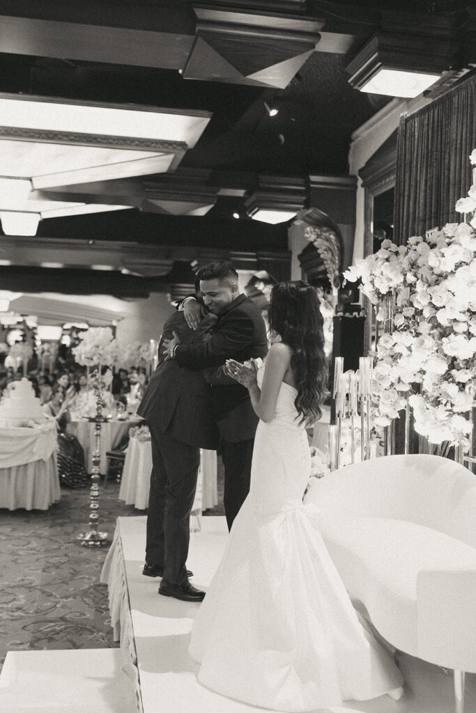 The groom hugs the best man on stage while the bride applauds during an emotional Catholic Indian wedding reception at Crest Hollow Country Club, a heartfelt celebration captured by Elsie Goodman Photography for an Indian wedding photo gallery.