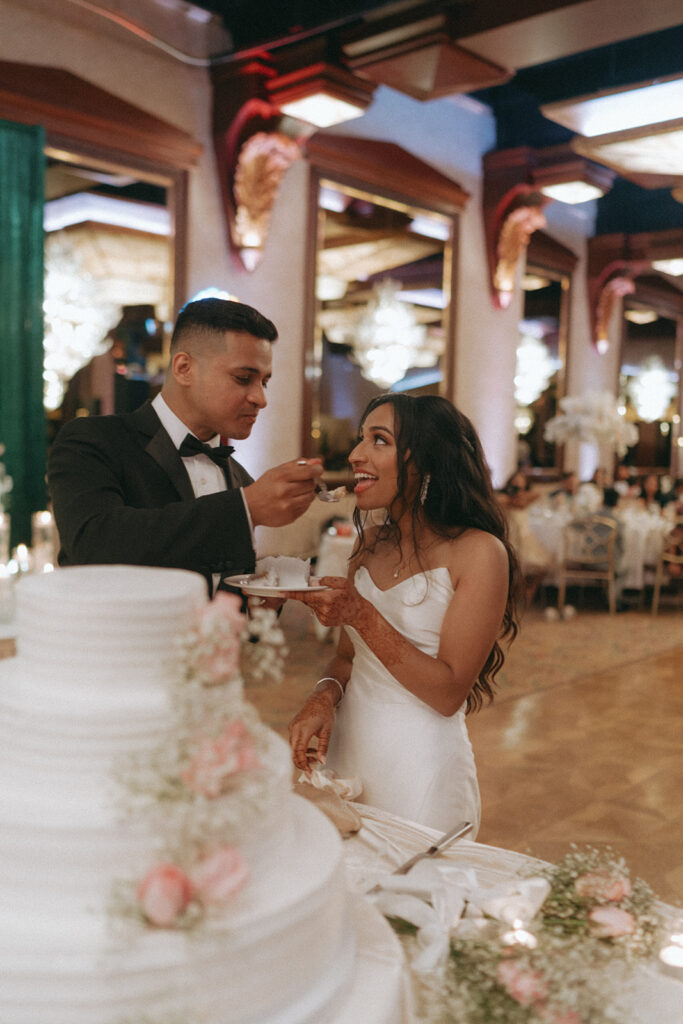 The groom feeds a bite of cake to the bride during their Catholic Indian wedding reception at Crest Hollow Country Club, a sweet and joyful moment documented by Elsie Goodman Photography in a Long Island wedding gallery.