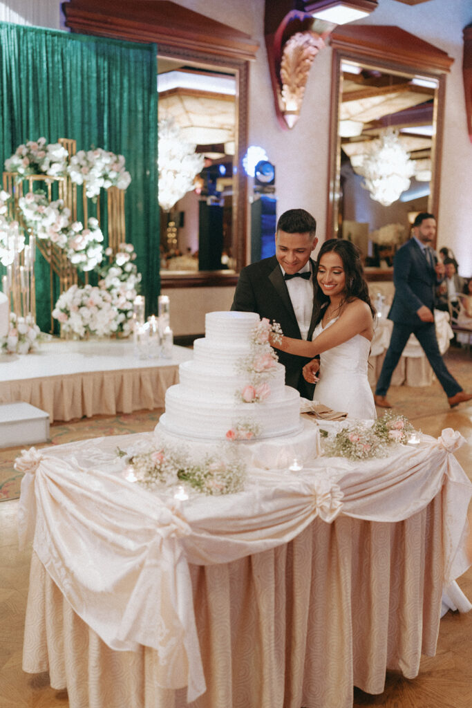 The newlyweds cut into their tiered wedding cake adorned with flowers during their elegant Catholic Indian wedding reception at Crest Hollow Country Club, a classic Long Island wedding scene captured by Elsie Goodman Photography.