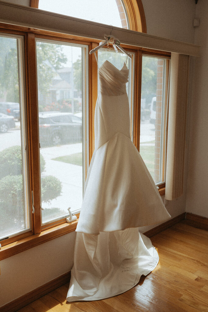 Bride’s white wedding gown hanging by sunlit window before Catholic Indian wedding ceremony — Elsie Goodman Photography.