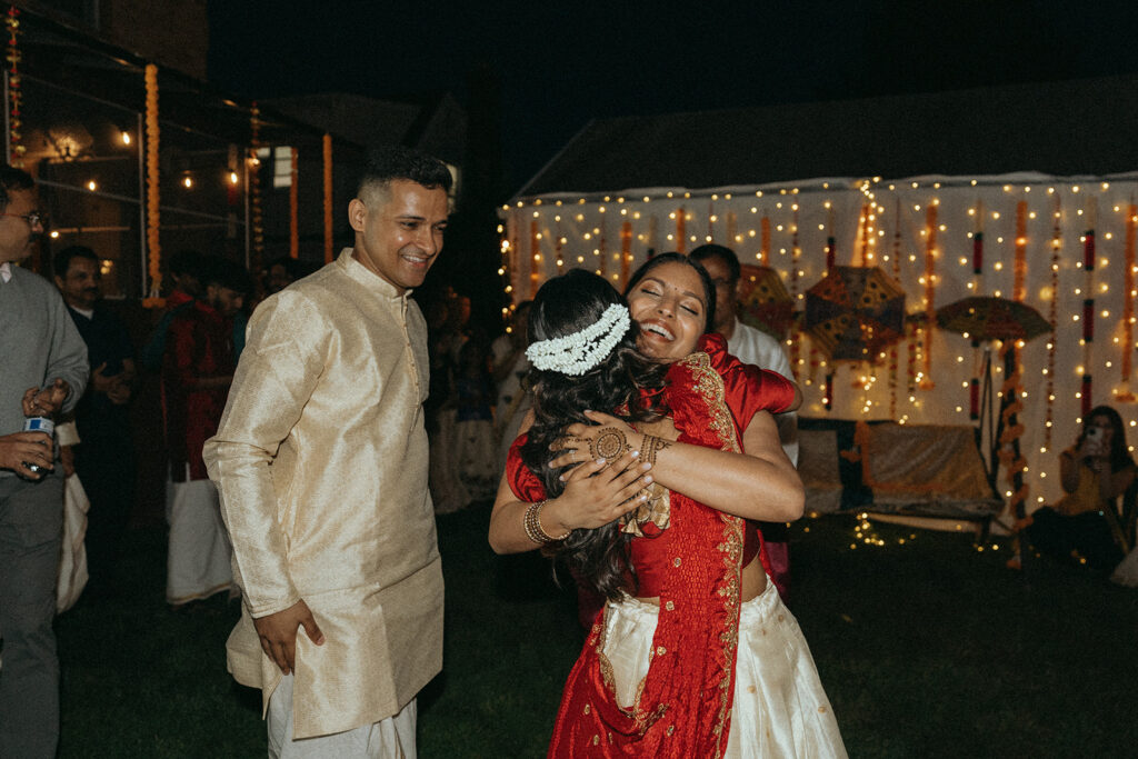 Bride hugging guest joyfully while groom smiles during Catholic Indian wedding welcome night — Elsie Goodman Photography.