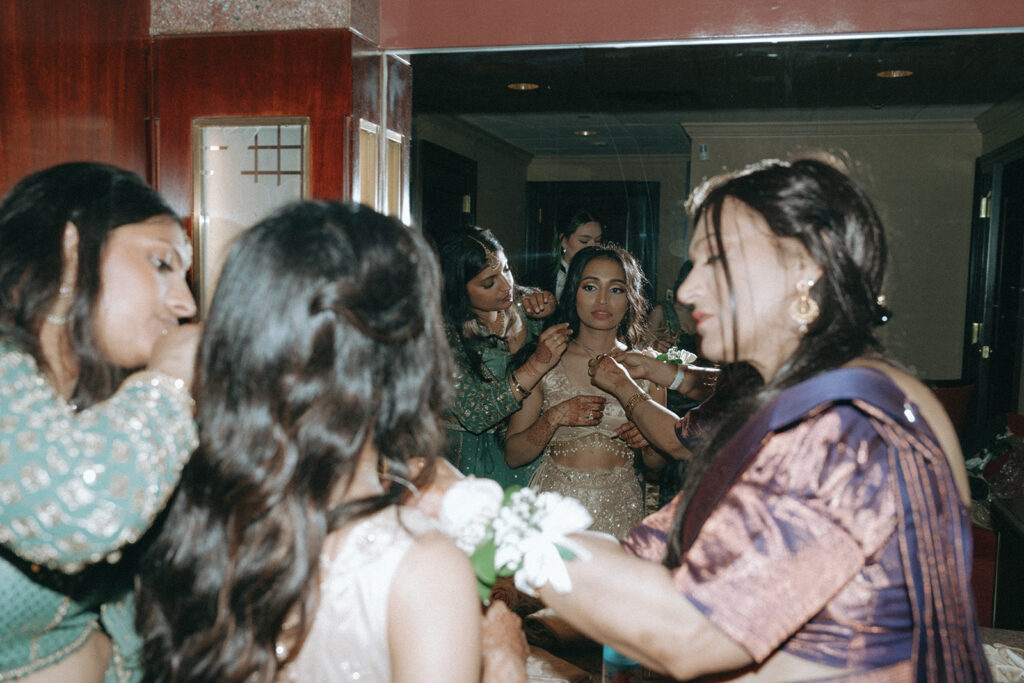 The bride getting final necklace adjustments from her bridesmaids and family while getting ready for her Indian Catholic wedding reception at Crest Hollow Country Club, captured by Elsie Goodman Photography.