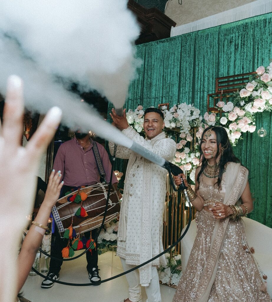 Bride and groom laughing joyfully while spraying smoke over the dance floor during their vibrant Catholic Indian wedding reception at Crest Hollow Country Club, beautifully captured by Elsie Goodman Photography for the Indian wedding photo gallery on Long Island.
