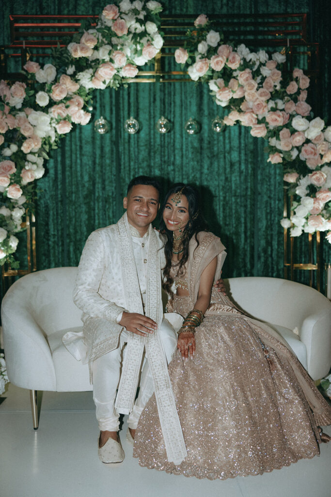 Bride and groom sitting together under a lush floral arch, smiling warmly during their Catholic Indian wedding reception at Crest Hollow Country Club, with the bride wearing a shimmering Indian catholic wedding dress, captured by Elsie Goodman Photography for the Indian wedding photo gallery.