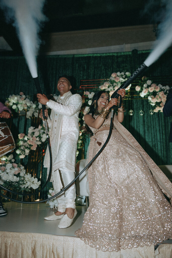 Bride and groom laughing as they shoot smoke cannons on stage during their lively Catholic Indian wedding reception at Crest Hollow Country Club, a top Indian wedding venue on Long Island, documented by Elsie Goodman Photography for the Indian wedding photo gallery.