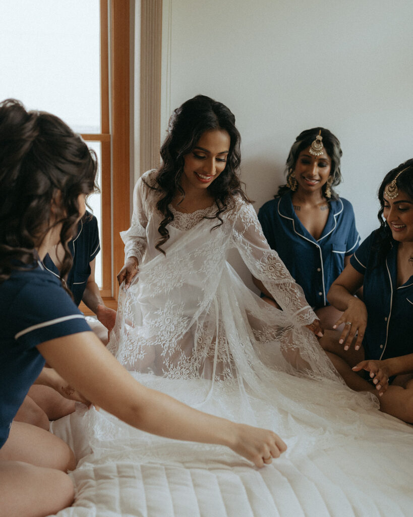 Bride laughing with bridesmaids while getting ready in white lace robe before Catholic Indian wedding — Elsie Goodman Photography.