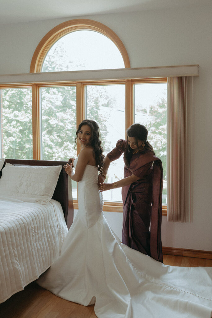 Alt Text: The bride smiling brightly as her mother buttons up her elegant white gown before the ceremony, blending Catholic Indian wedding traditions with modern bridal style, captured by Elsie Goodman Photography at a Long Island wedding venue.