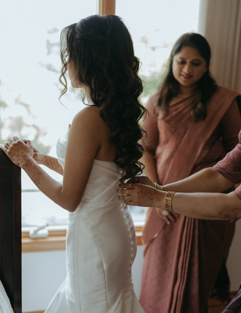 Bride getting help buttoning her wedding gown while standing by window before Catholic Indian wedding — Elsie Goodman Photography.