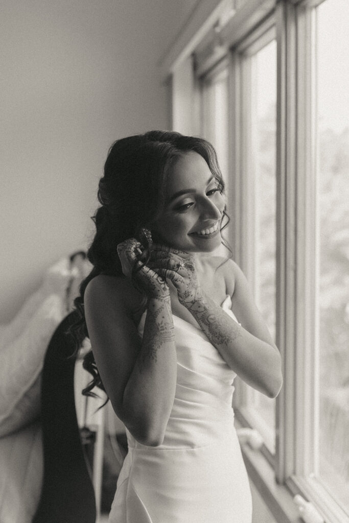 Bride smiling as she puts on earrings by the window, showing intricate mehndi designs on her hands, Catholic Indian wedding by Elsie Goodman Photography.