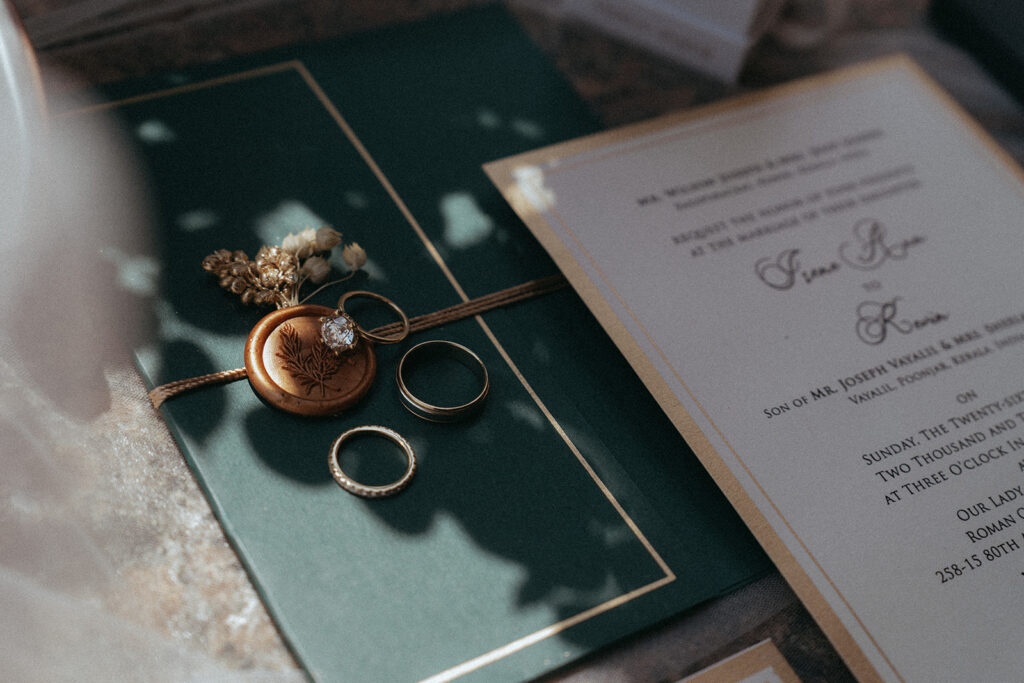 Flat lay of Catholic Indian wedding details including wedding rings, green invitation suite, and dried florals styled in soft natural light, by Elsie Goodman Photography.