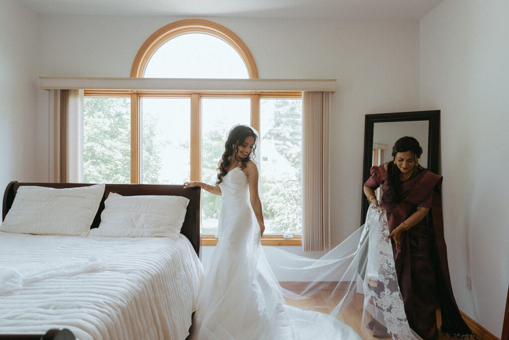 Bride getting ready for her Catholic Indian wedding as her mother adjusts her lace veil in a bright bedroom, captured by Elsie Goodman Photography.