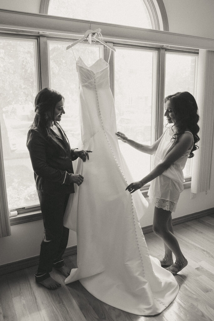 Bride and bridesmaid smiling and admiring the bride’s wedding dress hanging in front of a large arched window, photographed during Catholic Indian wedding preparations by Elsie Goodman Photography.