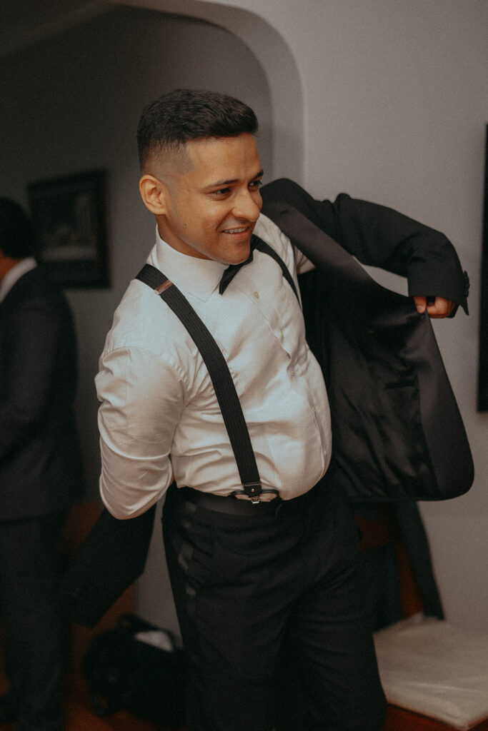 Groom smiling as he puts on his tuxedo jacket during Catholic Indian wedding getting ready photos, captured by Elsie Goodman Photography.