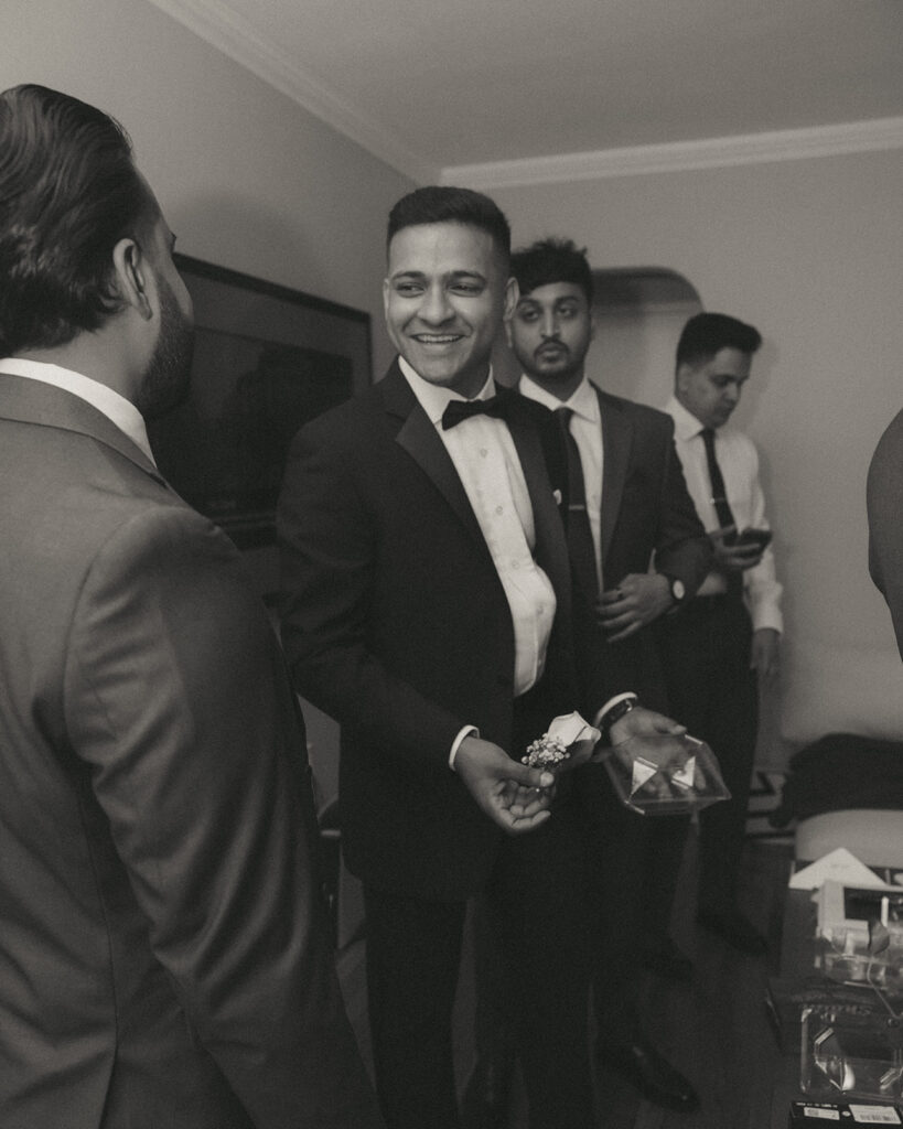 Groom smiling and holding boutonniere while chatting with groomsmen inside during Catholic Indian wedding morning preparations, photographed by Elsie Goodman Photography.