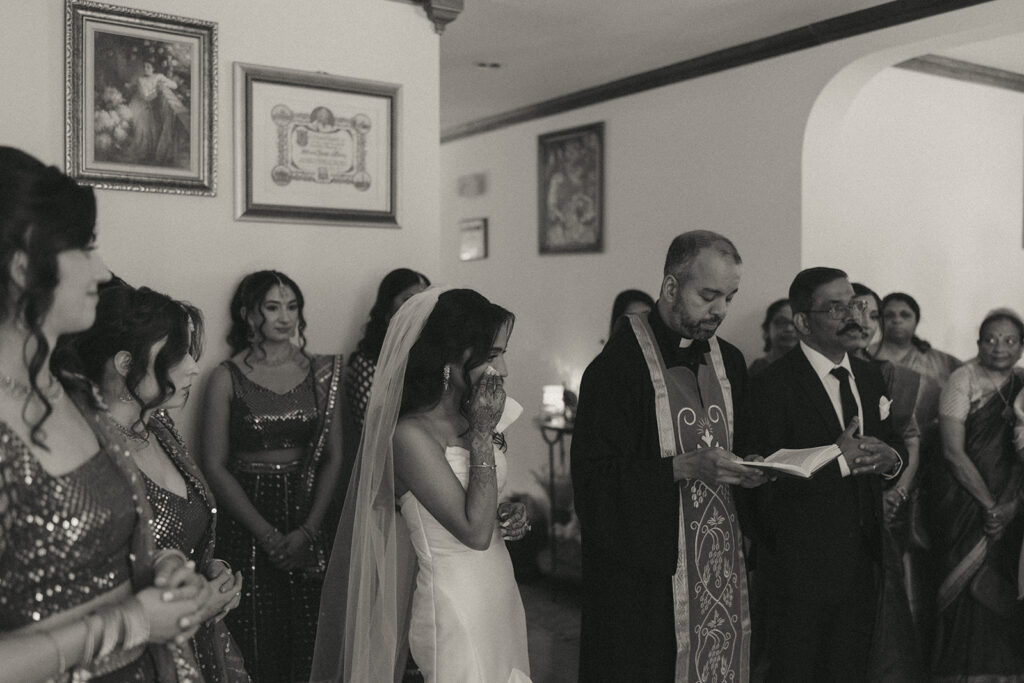 Bride wiping away tears during emotional Catholic Indian wedding family blessing, standing beside a priest and surrounded by her bridesmaids in glittering lehengas, captured by Elsie Goodman Photography.