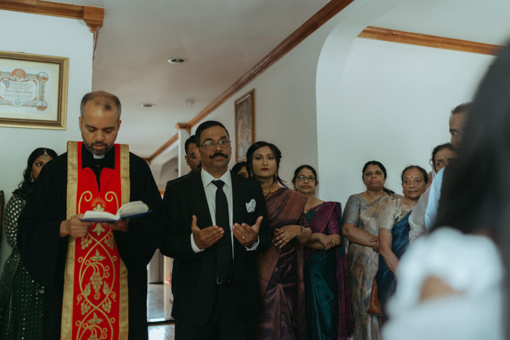 Catholic Indian wedding family prayer led by a priest as the bride's father prays beside him, with guests in colorful saris looking on, captured by Elsie Goodman Photography.