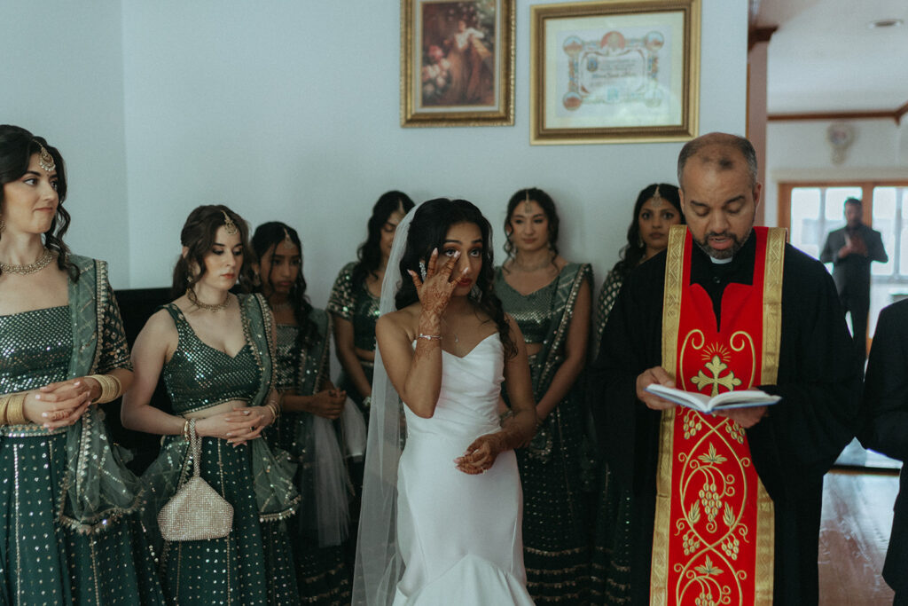 Bride wiping away tears during emotional Catholic Indian wedding family blessing, standing beside a priest and surrounded by her bridesmaids in glittering lehengas, captured by Elsie Goodman Photography.