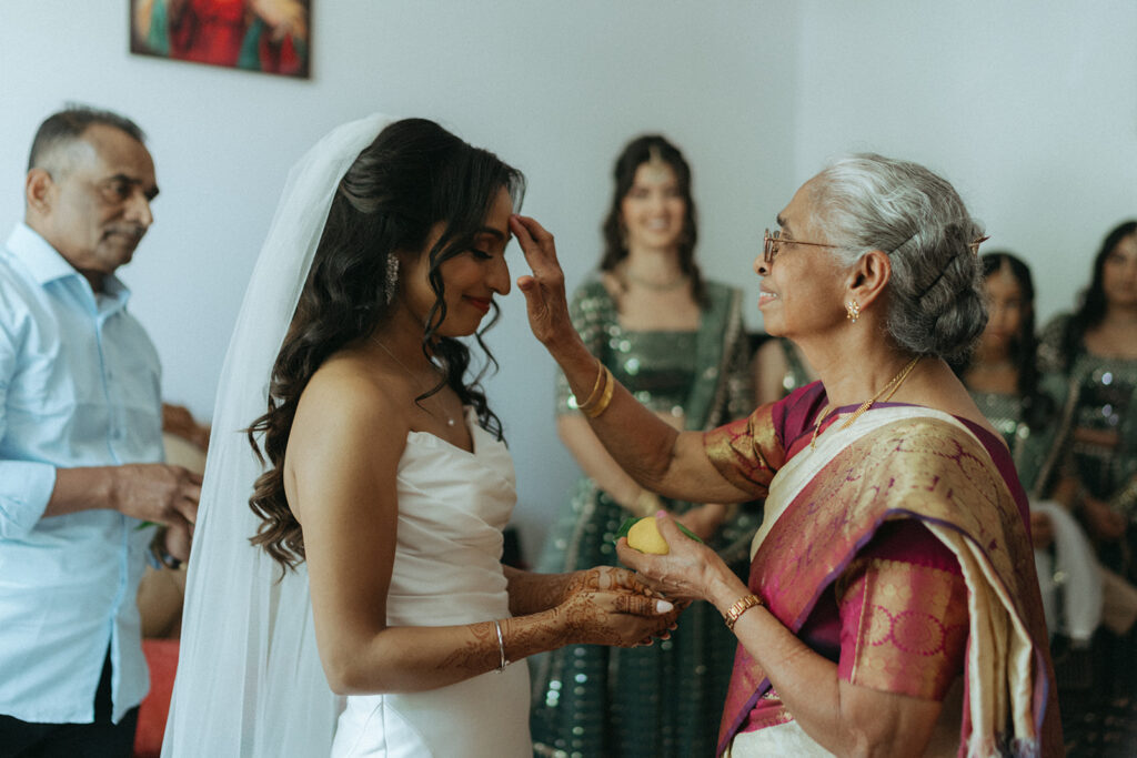 Grandmother performing a traditional blessing ceremony for the bride, who smiles with hands outstretched, surrounded by family at an Indian Catholic wedding, captured by Elsie Goodman Photography.