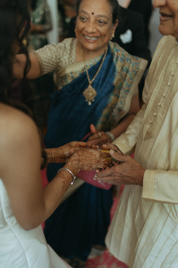 Alt Text: The bride's intricate mehndi-covered hands being held by an elder in a heartfelt moment during a Catholic Indian wedding ceremony, with family members in traditional attire smiling warmly, captured by Elsie Goodman Photography at a Long Island wedding venue.