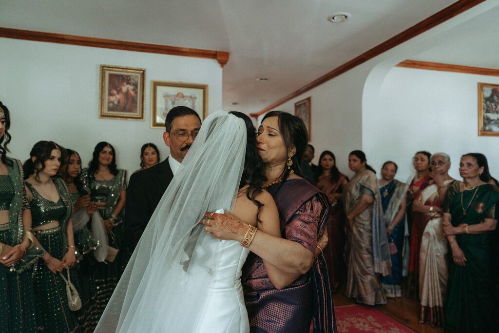 Bride sharing an emotional hug with her mother while family members and bridesmaids watch with tender smiles, a beautiful moment during Catholic Indian wedding preparations captured by Elsie Goodman Photography.