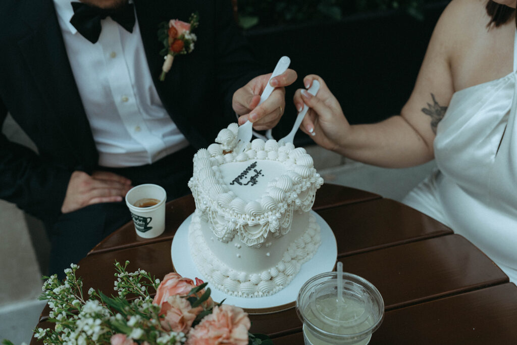 Close up of bride and groom with forks next to wedding cake with flowers, by Elsie Goodman
