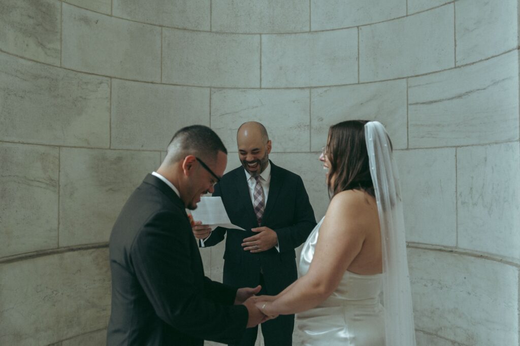 Couple stands in marble alcove with officiant during their NYPL wedding ceremony, by Elsie Goodman.