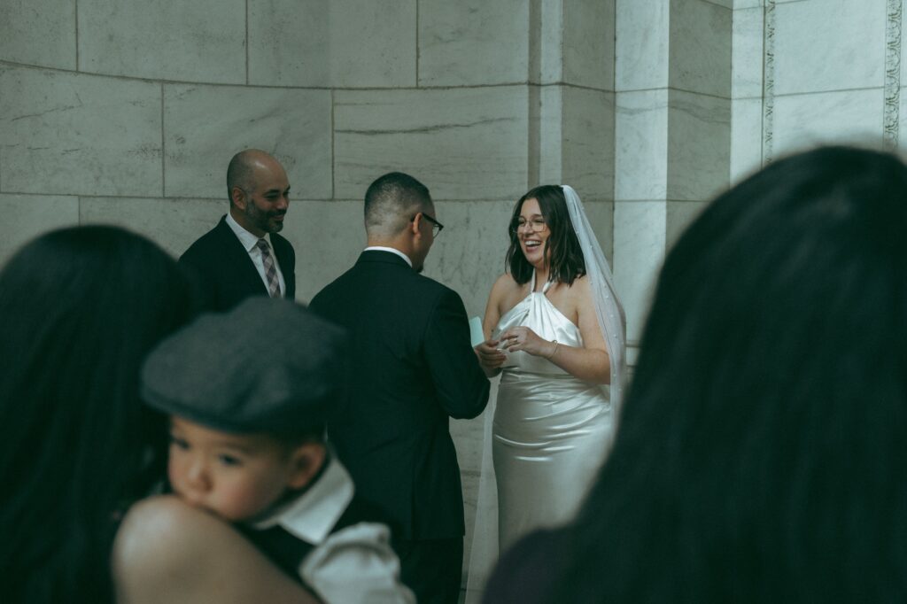 Bride laughs during NYPL wedding vows with guests in foreground, by Elsie Goodman Photography.