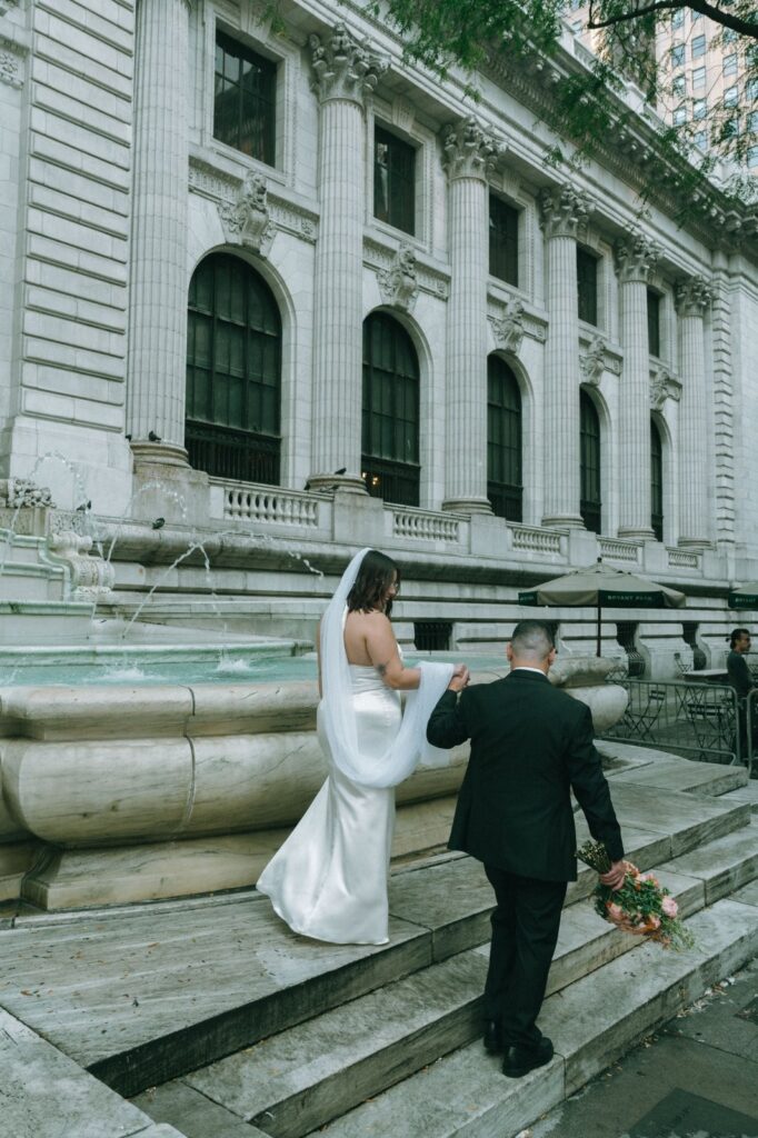 Bride and groom hold hands while walking in front of NYPL fountain after their wedding, by Elsie Goodman.
