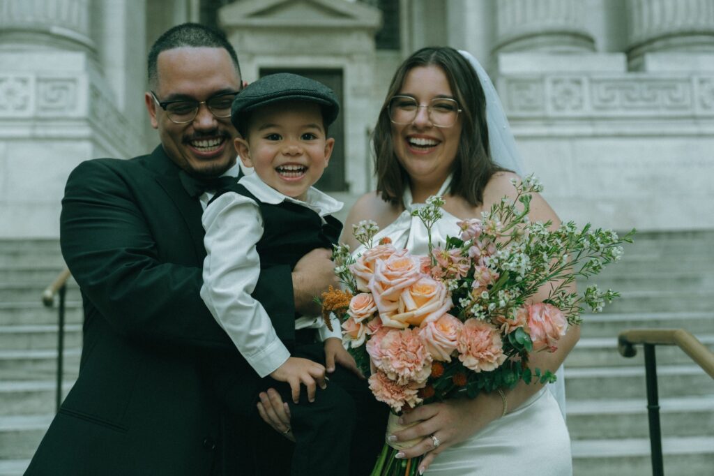 Bride and groom hold kid ring bearer outside of NYPL after wedding, by Elsie Goodman.