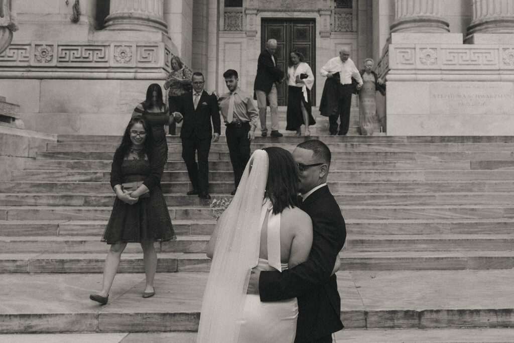 Black and white photo of couple kissing as guests join them after NYPL wedding ceremony, by Elsie Goodman.