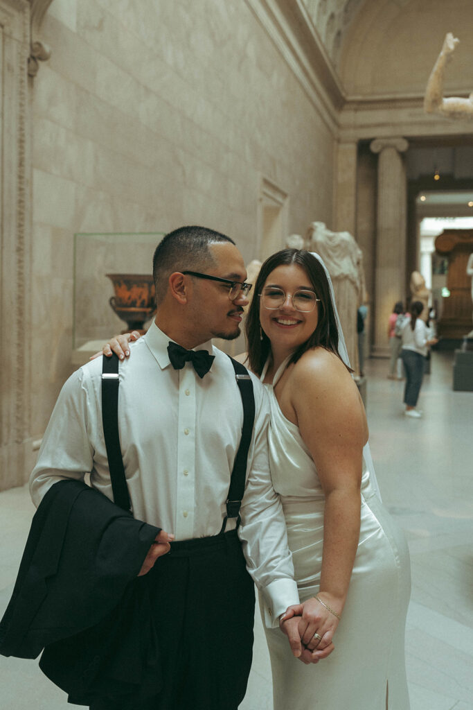 Bride smiling at camera while groom looks at her in museum after eloping at NYPL, by Elsie Goodman.