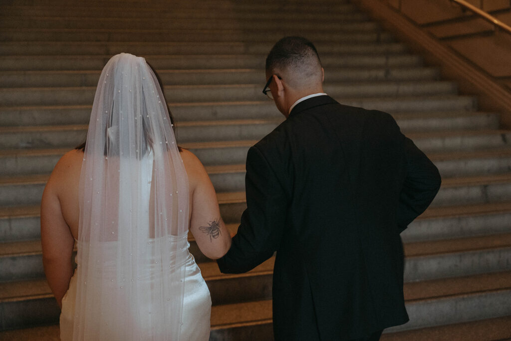Back view of bride and groom holding hands, walking up steps during New York Public Library elopement, by Elsie Goodman.