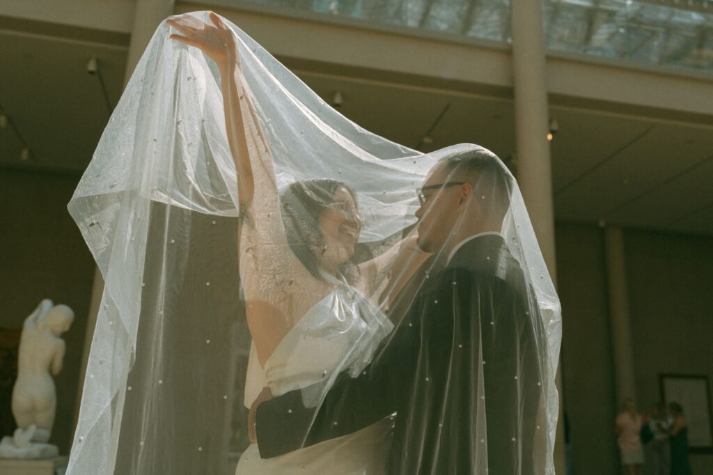 Bride lifts her veil over the couple as they laugh together inside a museum after their NYPL elopement, by Elsie Goodman.