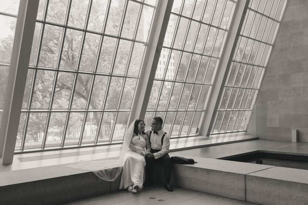 Black and white wedding photo of bride and groom sitting next to slanted windows at a museum, by Elsie Goodman.