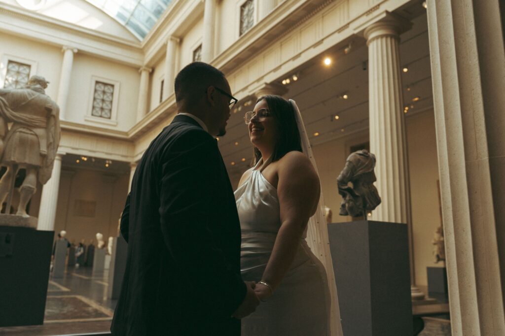 Bride and groom hold hands and smile at each other in the MET after their New York Public Library elopement, by Elsie Goodman.