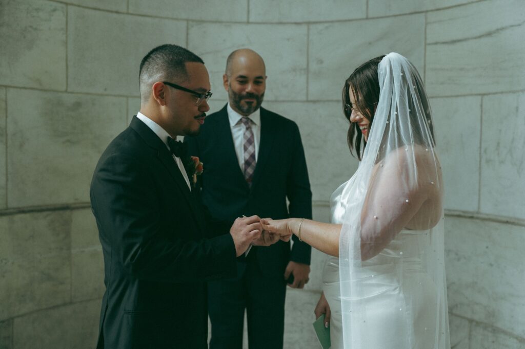 Groom places ring on bride’s hand at New York Public Library wedding, photographed by Elsie Goodman.