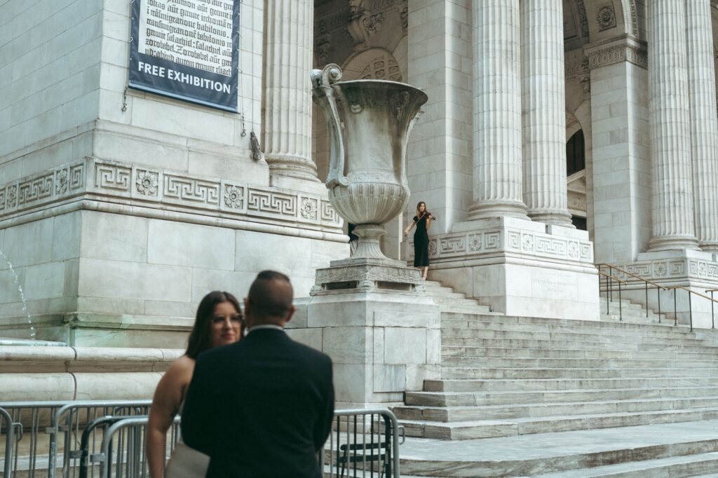 Violinist plays beside in background on library steps for an NYPL wedding with bride and fater out of focus in foreground, captured by Elsie Goodman.