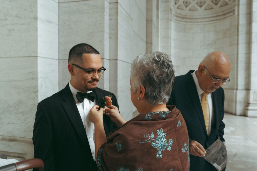 Woman pins red boutonniere to groom’s tux before New York Public Library wedding, photographed by Elsie Goodman.