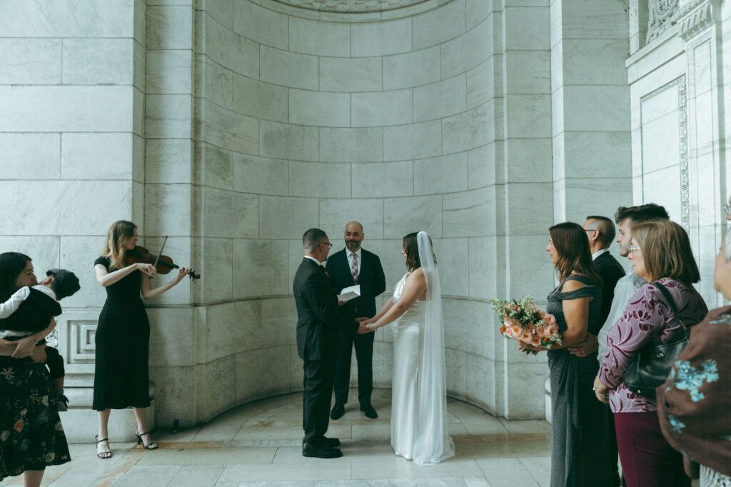 Bride and groom hold hands exchanging vows in a marble alcove as violinist plays and guests watch at NYPL wedding, photo by Elsie Goodman.