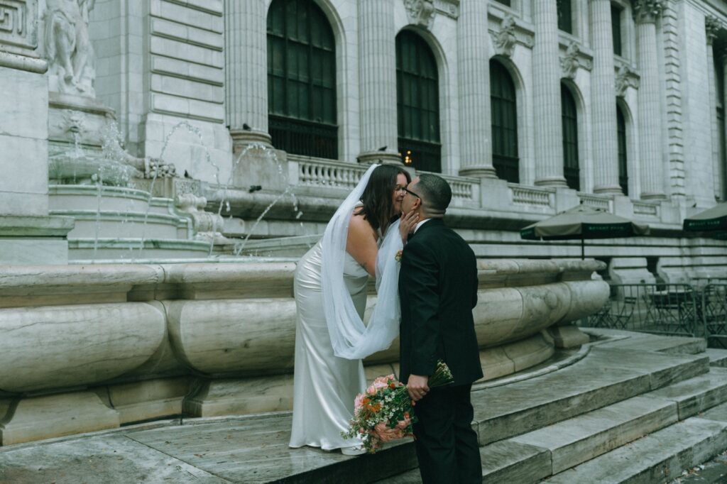 Bride leans down from fountain ledge to kiss groom holding bouquet at NYPL elopement, by Elsie Goodman