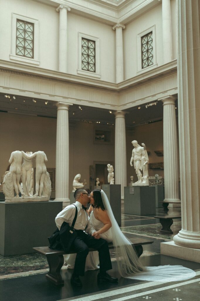 Bride and groom kiss on a bench surrounded by classical sculptures after NYPL elopement, captured by Elsie Goodman