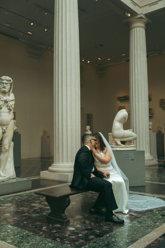 Bride smiles while touching groom’s face on a bench among sculptures after their wedding, by Elsie Goodman
