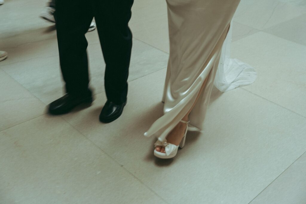 Close-up of bride’s white heels and groom’s black shoes walking on NYPL marble floor at elopement, by Elsie Goodman