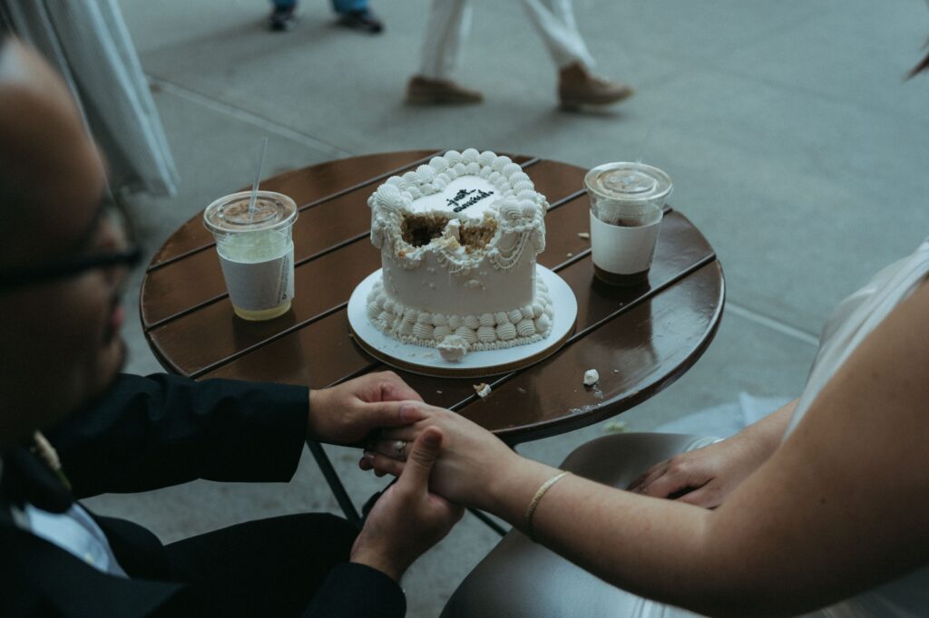 Bride and groom hold hand with Just Married wedding cake and coffee on a table in front of them, by Elsie Goodman.