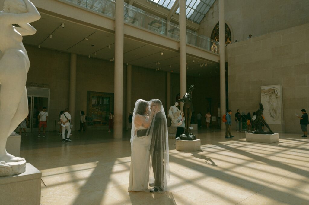 Bride and groom stand covered in bride's veil in a museum after their NYC elopement, by Elsie Goodman.
