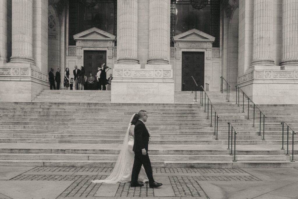Black and white photo of bride and her father walking into a NYPL wedding ceremony, captured by Elsie Goodman.