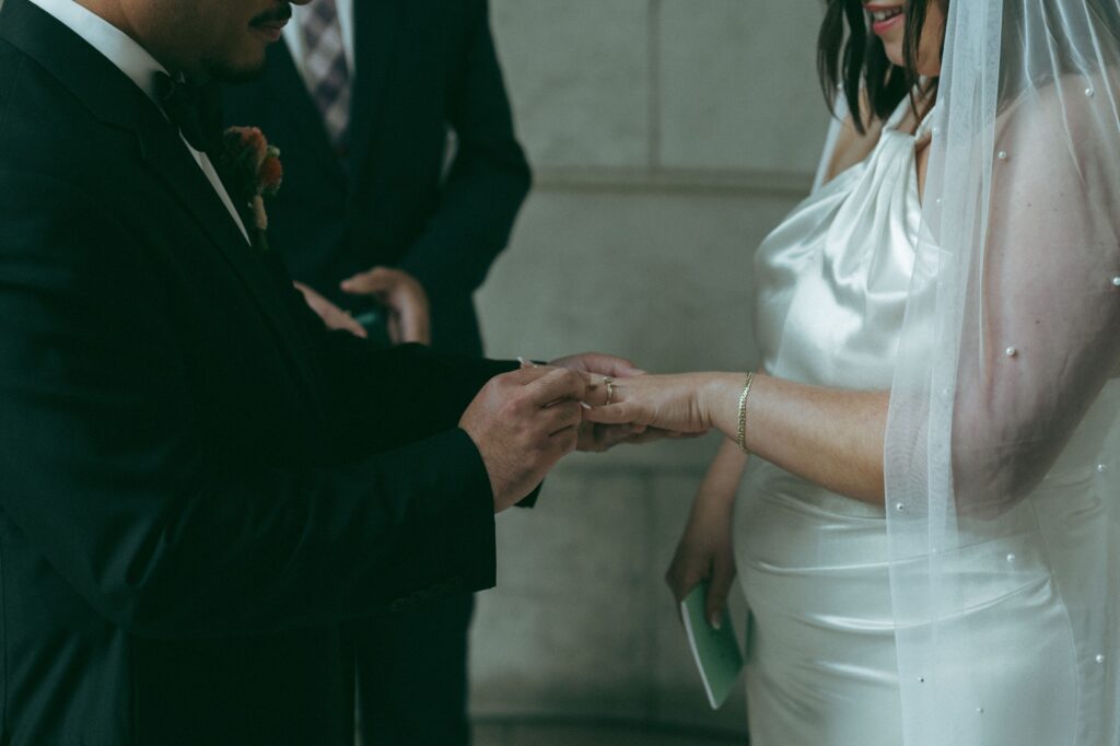 Close up of groom putting ring on bride's finger during NYPL wedding ceremony, shot by Elsie Goodman.