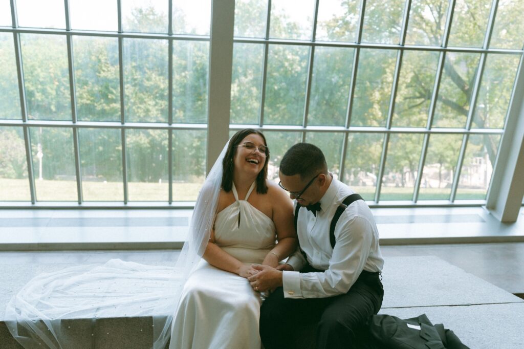 Bride and groom laugh, sitting in front of sunny window at the MET after eloping, captured by Elsie Goodman.