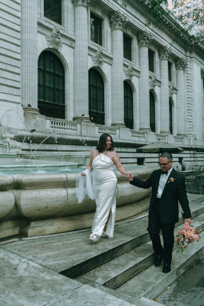 Bride and groom walk hand in hand on fountain steps outside of NYPL after eloping, shot by Elsie Goodman.